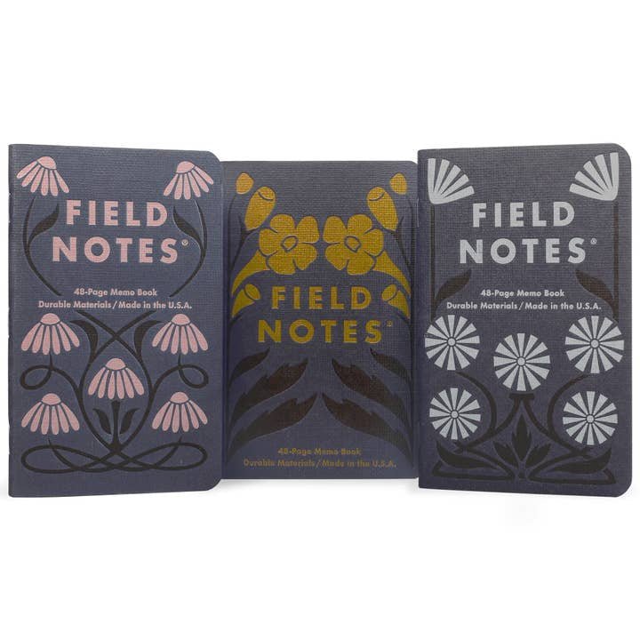 The Field Notes Fall Flowers 3-Pack includes three 48-page memo books with early-20th-century-inspired botanical covers, featuring pink, yellow, and white fall flowers on dark backgrounds, standing upright side by side.