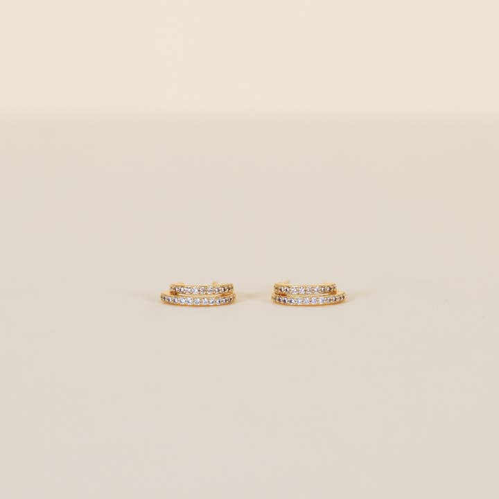 A pair of Double Champagne Hoop earrings by JaxKelly, featuring small gold double hoops adorned with two rows of shimmering pave stones, displayed against a plain beige background.
