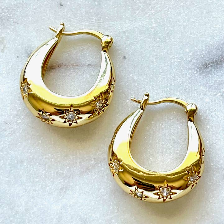 The Aviva Earrings by Jessica Matrasko Jewelry are gold filled huggie earrings adorned with star-shaped CZ diamond accents, elegantly displayed on a light marble surface. Hypoallergenic and perfect for everyday elegance, these earrings bring a subtle sparkle to any look.
