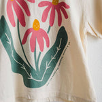The Sow the Seeds ivory boxy crop tee from Ramble & Company features a graphic of three pink coneflowers with yellow centers and green leaves, making it the perfect casual shirt for any garden enthusiast.