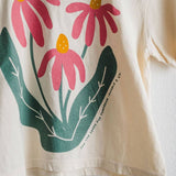 The Sow the Seeds ivory boxy crop tee from Ramble & Company features a graphic of three pink coneflowers with yellow centers and green leaves, making it the perfect casual shirt for any garden enthusiast.