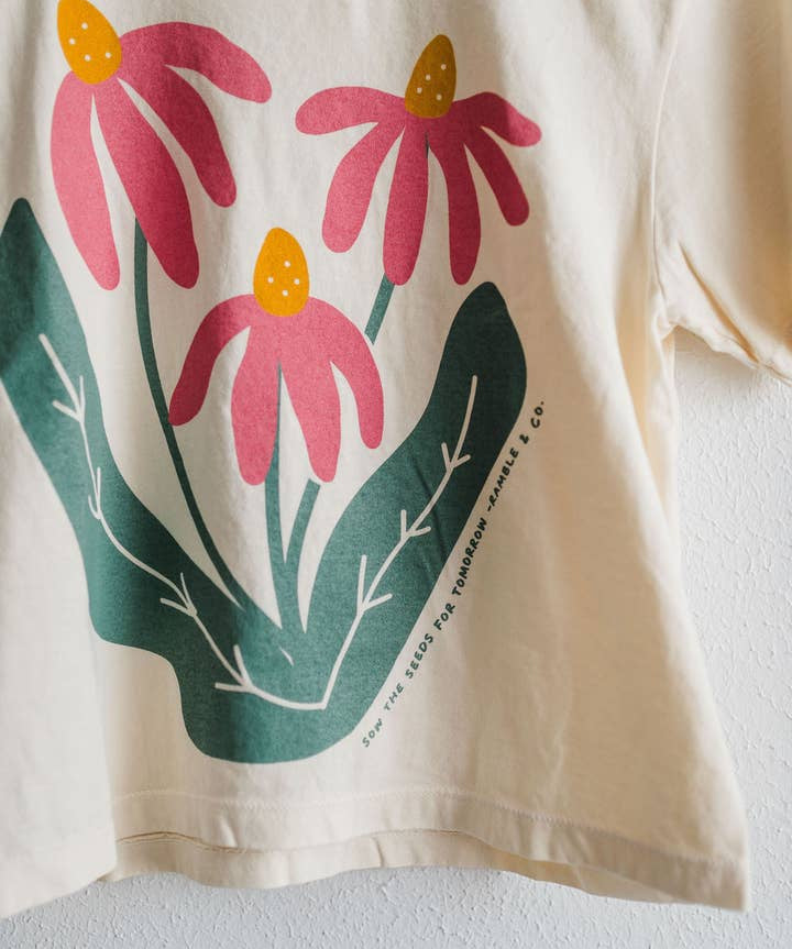 The Sow the Seeds ivory boxy crop tee from Ramble & Company features a graphic of three pink coneflowers with yellow centers and green leaves, making it the perfect casual shirt for any garden enthusiast.