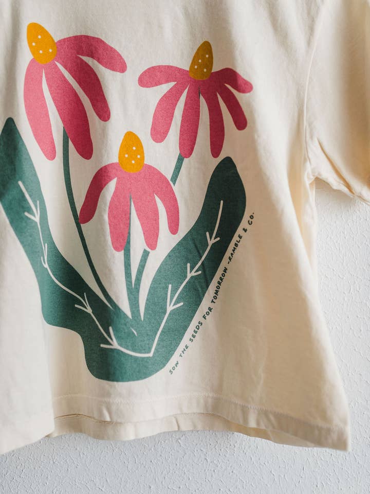 The Sow the Seeds ivory boxy crop tee from Ramble & Company features a graphic of three pink coneflowers with yellow centers and green leaves, making it the perfect casual shirt for any garden enthusiast.