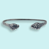 Discover the Silver Snake Cuff Bracelet by Ornamental Things, an elegant and bold accessory featuring two eye-catching snakehead designs facing each other. This adjustable silver bracelet stands out against a light blue background, seamlessly blending sophistication with statement style.