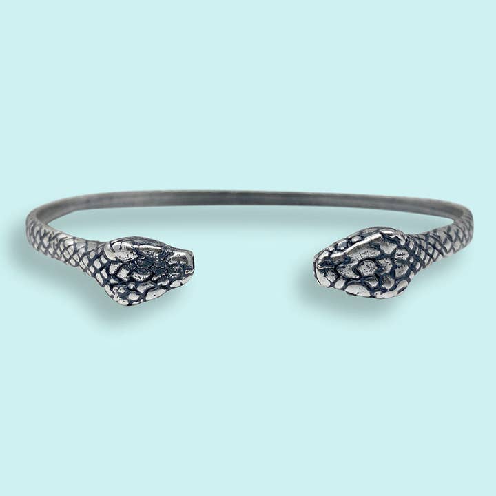 Discover the Silver Snake Cuff Bracelet by Ornamental Things, an elegant and bold accessory featuring two eye-catching snakehead designs facing each other. This adjustable silver bracelet stands out against a light blue background, seamlessly blending sophistication with statement style.