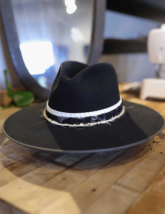 The Easy Drifter Felt Hat in Black Valley Crease, featuring a wide brim and white decorative band, rests on a wooden surface with a mirror and sewing machine in the background.