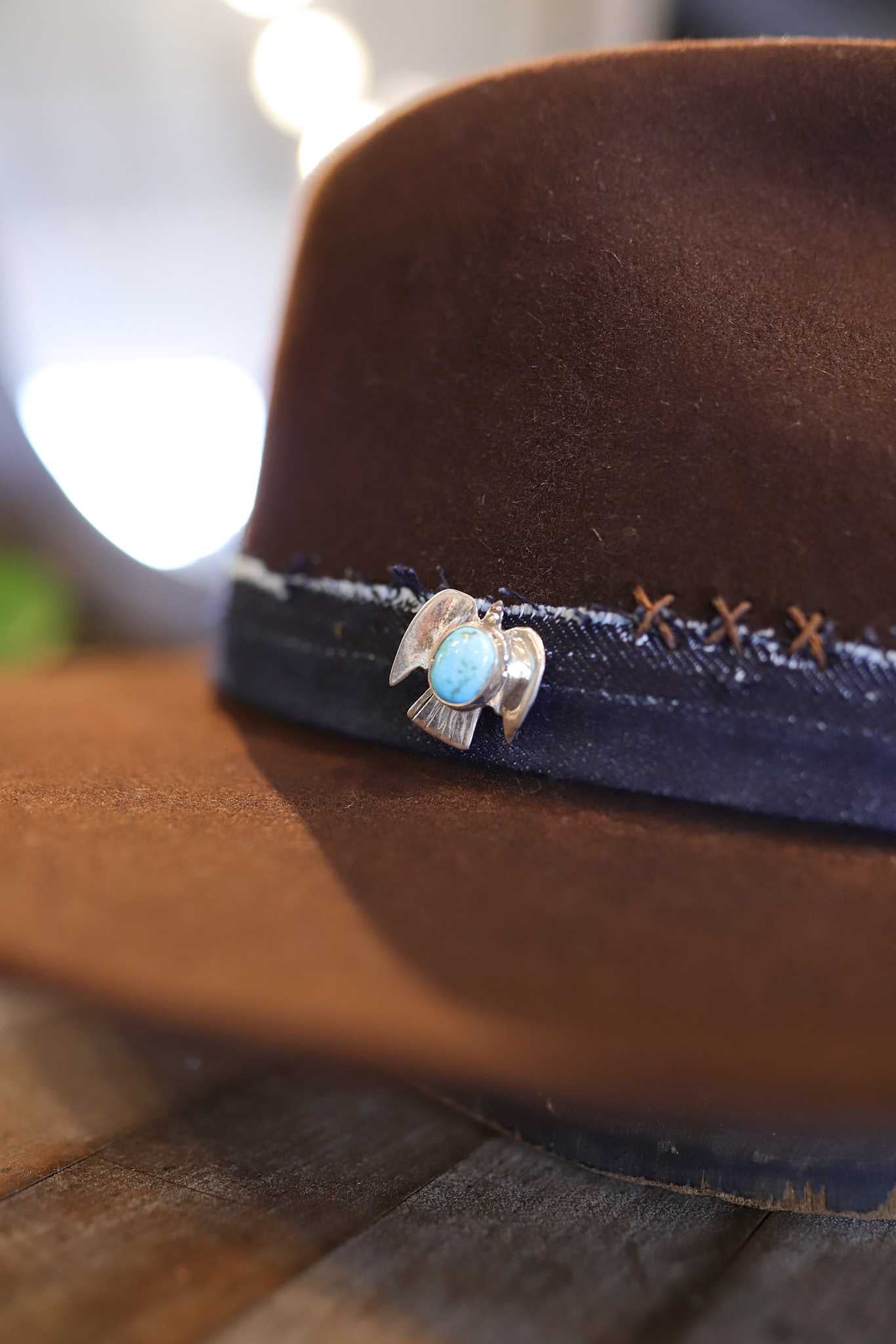A close-up of the Easy Drifter Felt Hat in Chocolate Front Pinch, featuring a sterling silver turquoise hat pin on a black band, resting on a wooden surface.