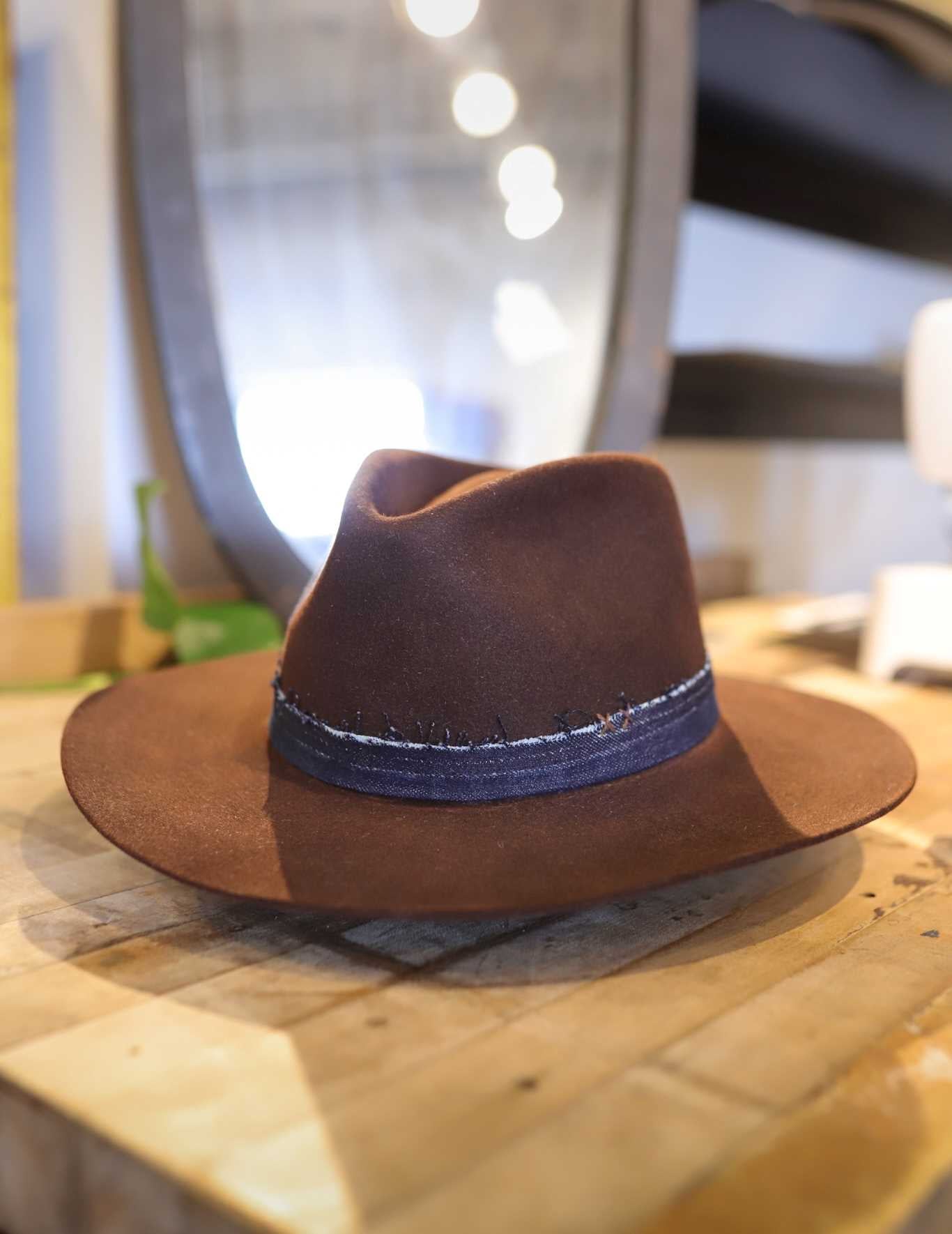 The Easy Drifter Felt Hat | Chocolate Front Pinch rests on a wooden table, featuring a dark blue band and a sterling silver turquoise pin; a mirror and blurred elements accent the background.