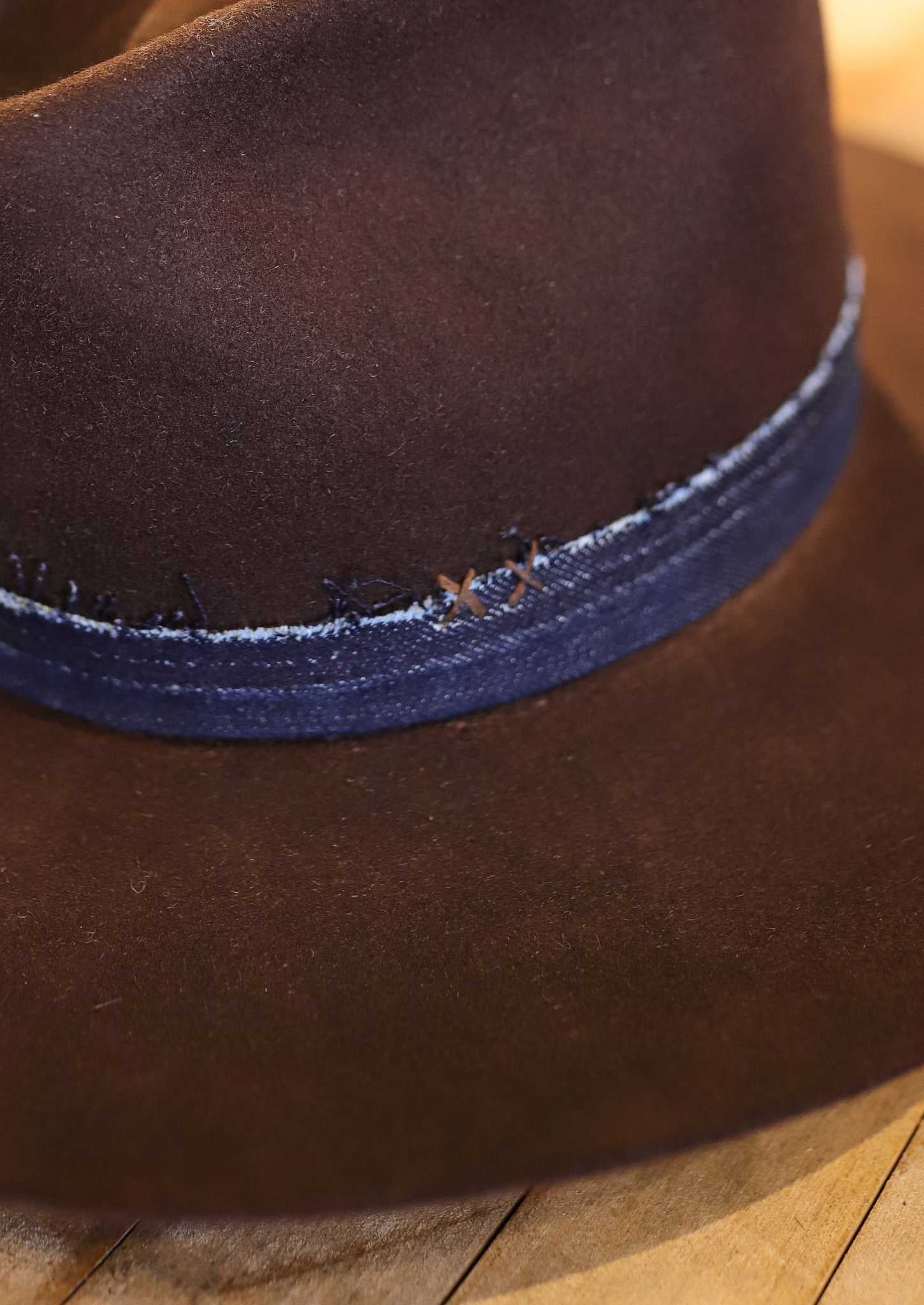 The Easy Drifter Felt Hat in Chocolate features a front pinch fedora style with a blue stitched, frayed fabric band, shown resting on wood.