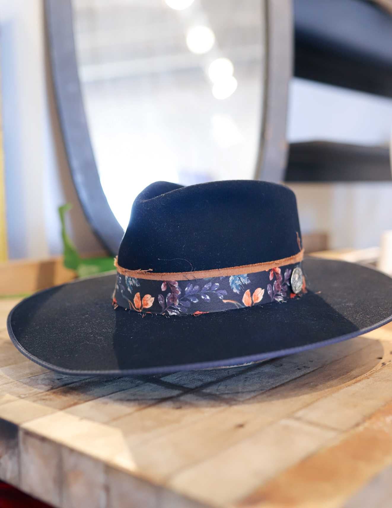 The Easy Drifter Felt Hat in Navy with a front pinch style rests on a wooden surface, accented by a floral hat band and leather trim, with a blurred mirror in the background.