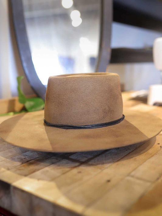 The Easy Drifter Felt Hat | Pecan Stove Pipe features a light brown, handmade design with a thin black band, shown resting on wood with a blurred mirror and green plant in the background.