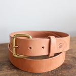 The Field Duty Belt by Ballad of the Bird Dog, crafted from tan Wickett & Craig harness leather with a brass buckle, is neatly coiled on a wooden surface.