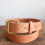 The Field Duty Belt by Ballad of the Bird Dog, made with tan Wickett & Craig harness leather and a brass buckle, is coiled on a wooden surface.