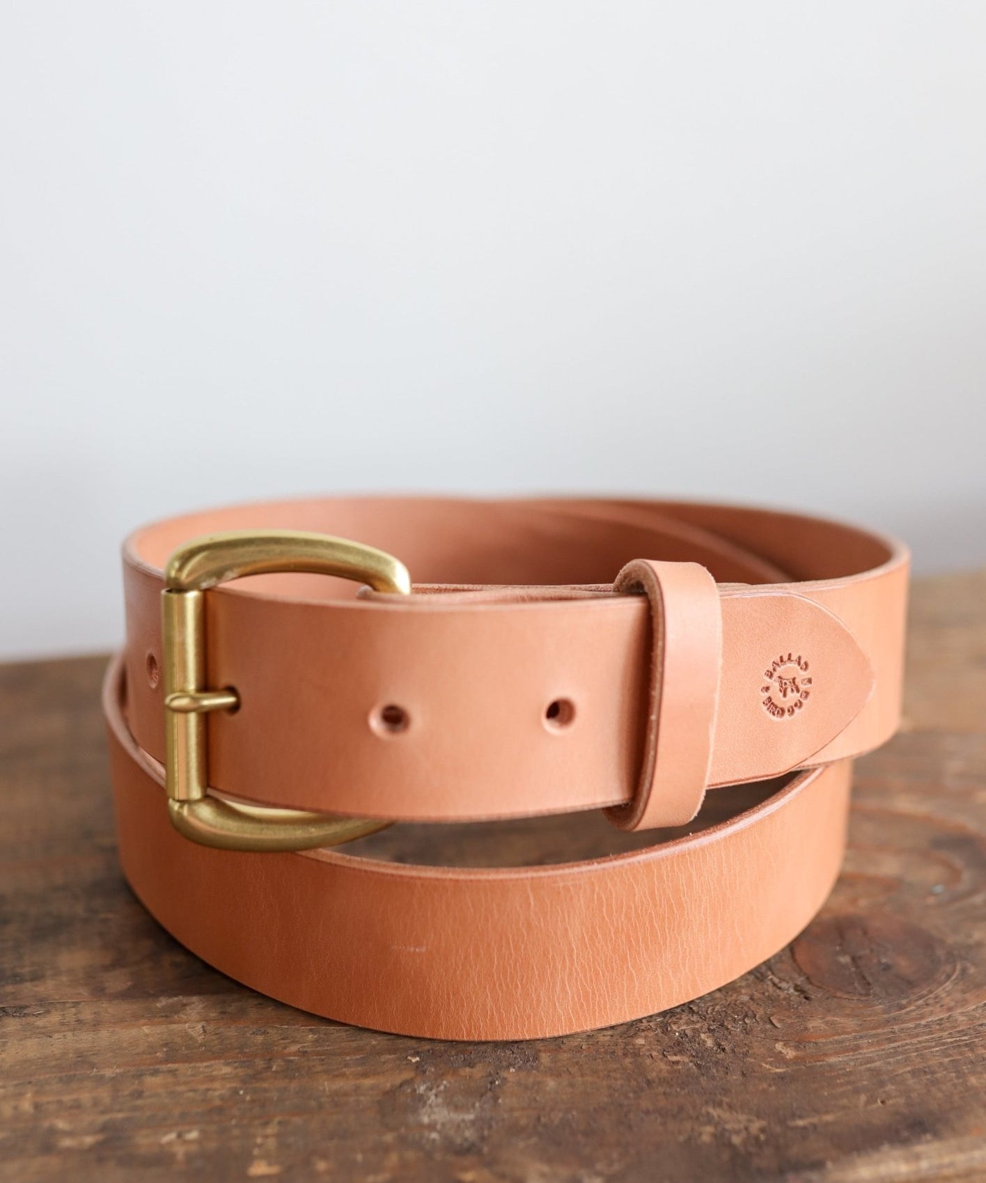 The Field Duty Belt by Ballad of the Bird Dog, made with tan Wickett & Craig harness leather and a brass buckle, is coiled on a wooden surface.