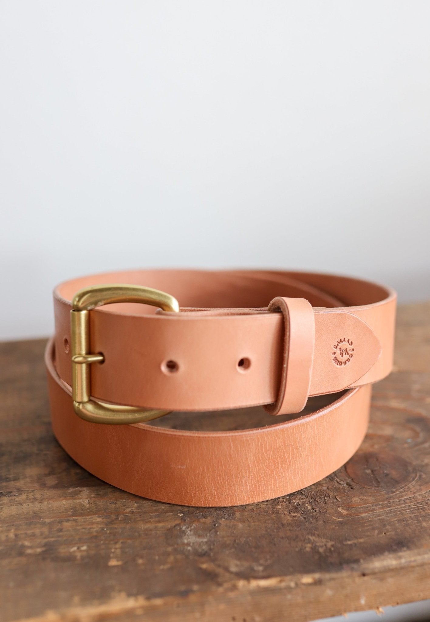 The Field Duty Belt by Ballad of the Bird Dog, made with tan Wickett & Craig harness leather and a brass buckle, is coiled on a wooden surface.