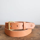 The Field Duty Belt by Ballad of the Bird Dog, made from vegetable tanned tan harness leather with a gold buckle and embossed logo near the buckle, is shown coiled on a wooden surface.
