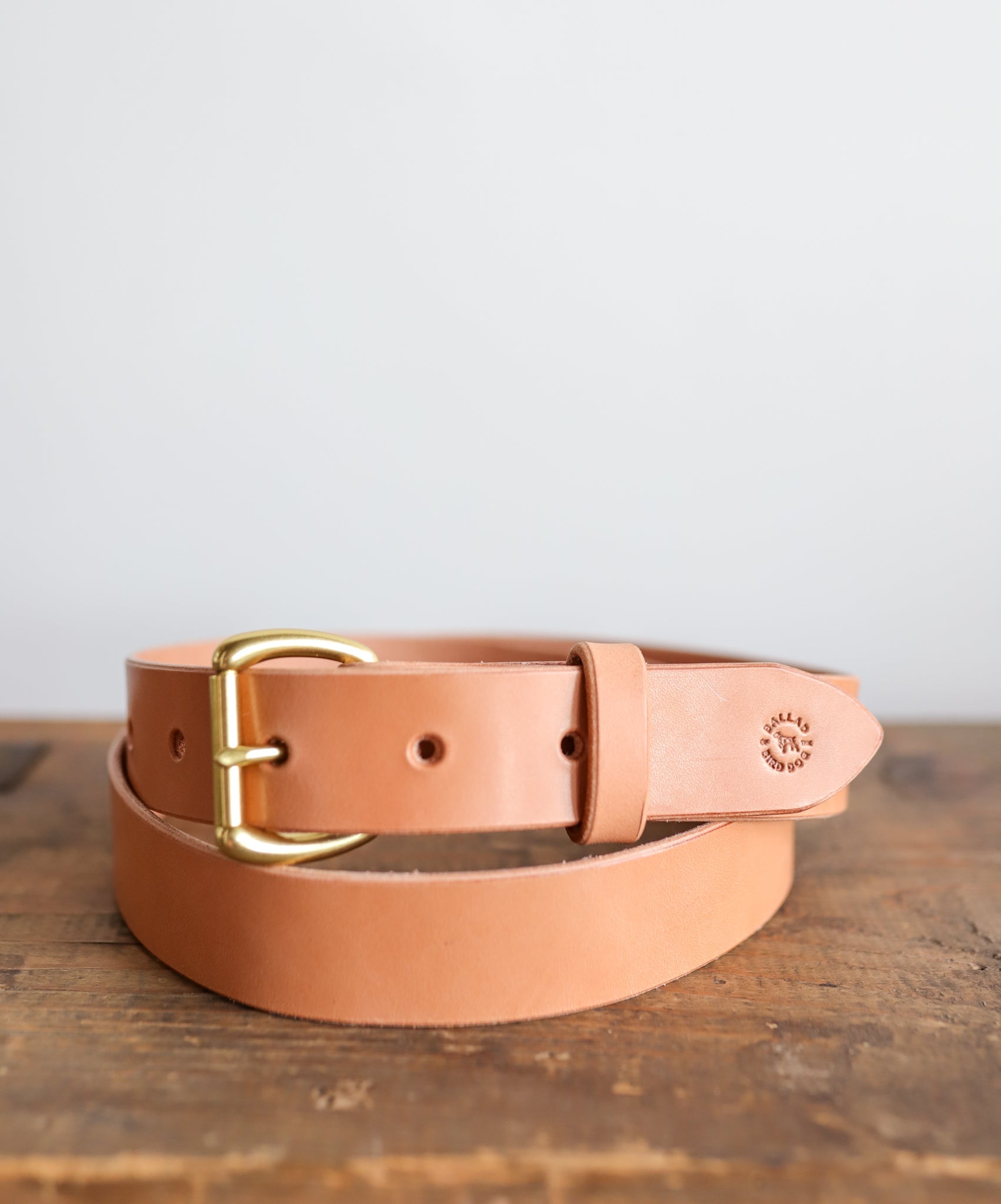 The Field Duty Belt by Ballad of the Bird Dog, made from vegetable tanned tan harness leather with a gold buckle and embossed logo near the buckle, is shown coiled on a wooden surface.
