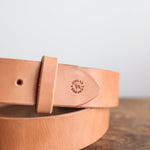 The Field Duty Belt by Ballad of the Bird Dog, made from tan Wickett & Craig vegetable tanned harness leather and stamped with a “DALLAS BEND GOODS” logo, rests on a wooden surface.