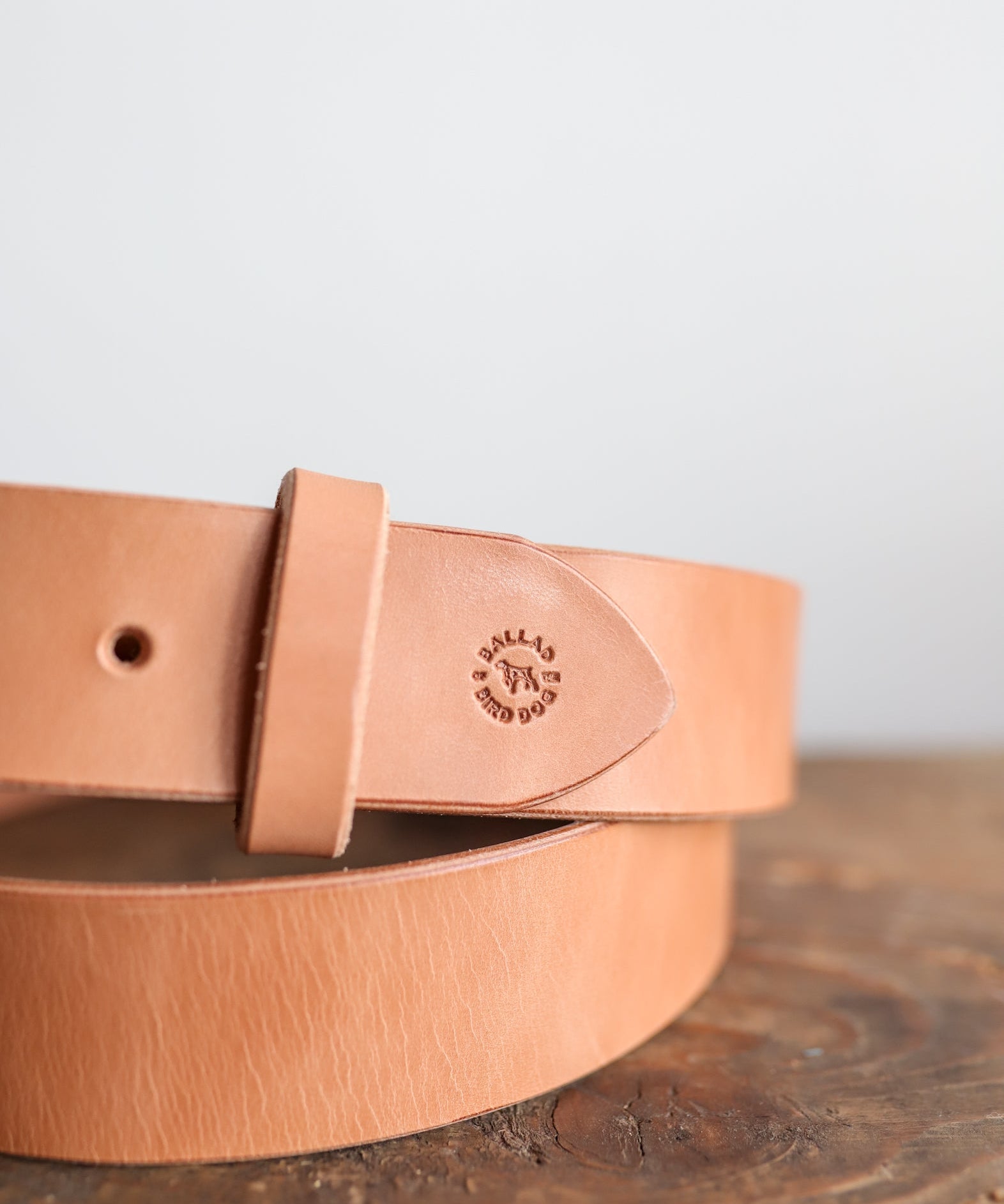 The Field Duty Belt by Ballad of the Bird Dog, crafted from premium tan harness leather, features a loop and an embossed logo. Made from Wickett & Craig vegetable tanned leather, it rests on a wooden surface against a plain background.