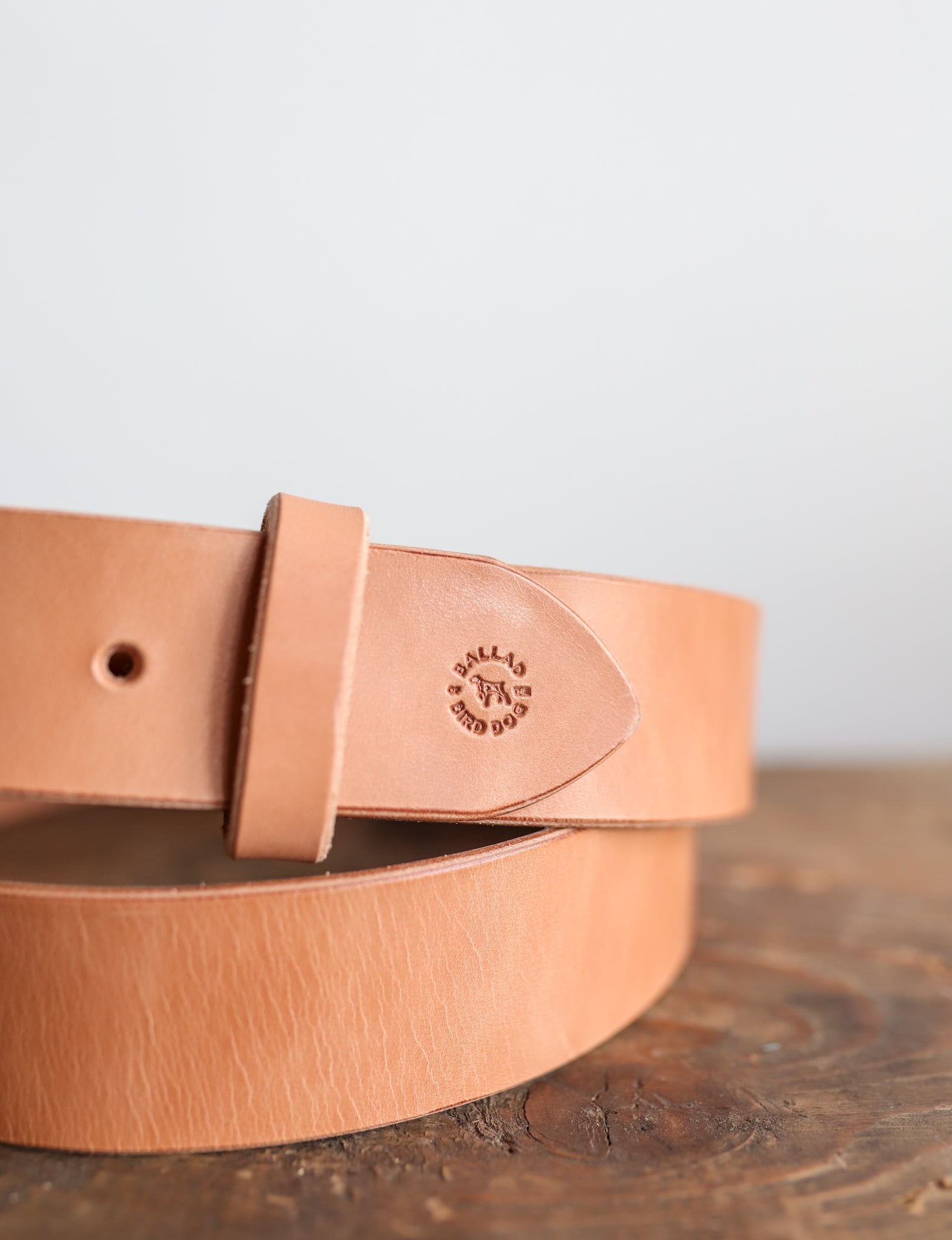 The Field Duty Belt by Ballad of the Bird Dog, crafted from premium tan harness leather, features a loop and an embossed logo. Made from Wickett & Craig vegetable tanned leather, it rests on a wooden surface against a plain background.