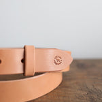 A tan Field Duty Belt by Ballad of the Bird Dog, made from vegetable-tanned harness leather with a rounded tip and "CARRELLO HAND BORN" logo, rests on a wooden surface.