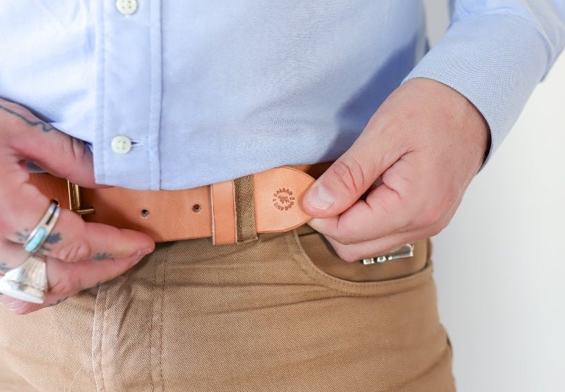 A person in a light blue shirt and tan pants fastens the Ballad of the Bird Dog Field Duty Belt, showing its subtle stamped logo, while silver rings adorn their left hand.
