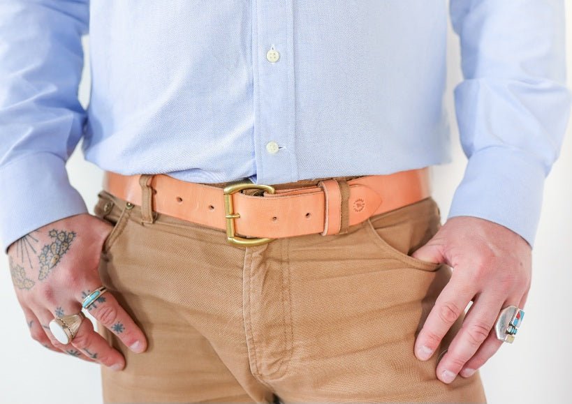 A person wearing a light blue button-up shirt, tan pants, and the Field Duty Belt by Ballad of the Bird Dog with a gold buckle, along with several rings on both hands.