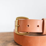 The Field Duty Belt by Ballad of the Bird Dog, crafted from tan Wickett & Craig harness leather with a gold buckle, is displayed on a wooden surface against a plain light background.