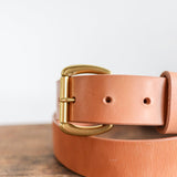 A close-up of the Ballad of the Bird Dog Field Duty Belt, featuring a gold buckle and vegetable tanned Wickett & Craig harness leather, shown resting on wood against a plain white background.