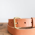The Field Duty Belt by Ballad of the Bird Dog features Wickett & Craig harness leather in tan and a gold buckle, crafted from vegetable-tanned leather. Displayed on wood with a plain background.