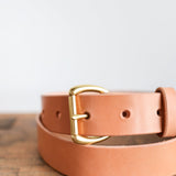 The Field Duty Belt by Ballad of the Bird Dog features tan Wickett & Craig harness leather and a gold D-shaped buckle, shown coiled on wood with a light background.