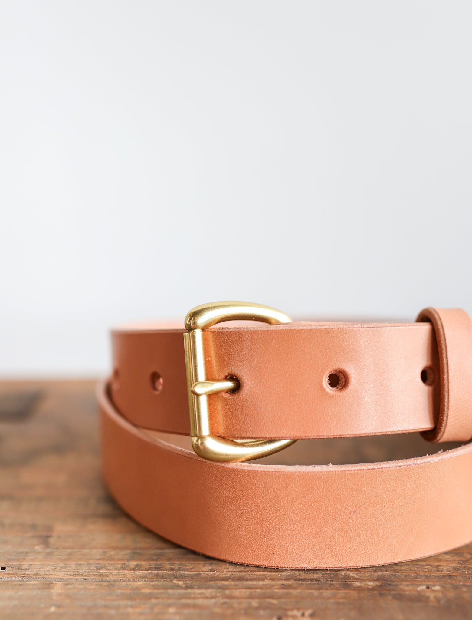The Field Duty Belt by Ballad of the Bird Dog features tan Wickett & Craig harness leather and a gold D-shaped buckle, shown coiled on wood with a light background.