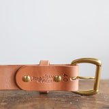 The Field Duty Belt by Ballad of the Bird Dog features light tan Wickett & Craig veg-tan harness leather, brass buckle and rivets, and "Brass Tacks" embossing, shown resting on a wooden surface.
