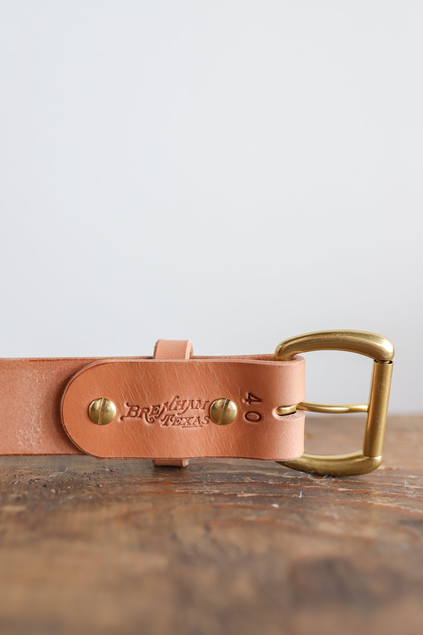 The Field Duty Belt by Ballad of the Bird Dog features light tan Wickett & Craig veg-tan harness leather, brass buckle and rivets, and "Brass Tacks" embossing, shown resting on a wooden surface.