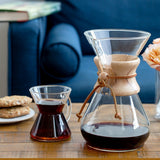 A Chemex Filter Drip Coffeemaker (CM-10A) and a small glass cup of coffee sit on a wooden table with cookies, with a blue couch in the background, highlighting Chemex's classic 50 oz design and timeless style.