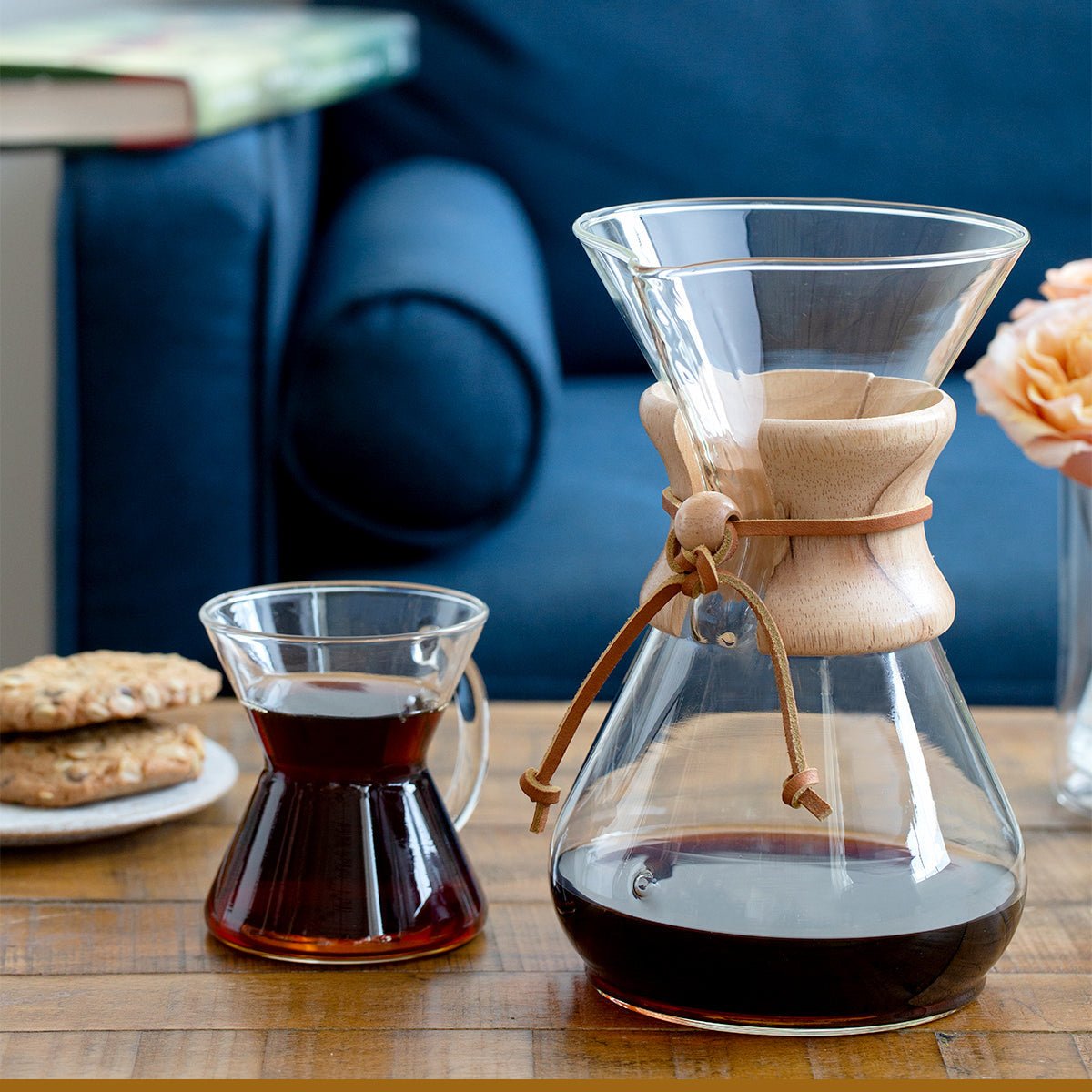 A Chemex Filter Drip Coffeemaker (CM-10A) and glass mug with coffee rest on a wooden table beside cookies and a peach rose, with a blue couch in the background—a cozy scene for enjoying this ten-cup Chemex coffee maker.