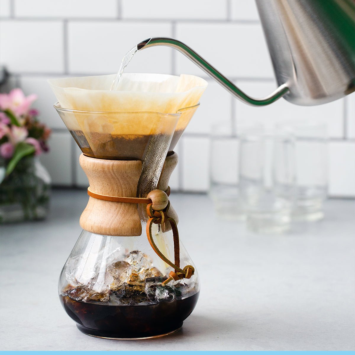A hand pours hot water from a kettle into a Chemex Filter Drip Coffeemaker | CM-6A with a paper filter and ice on a kitchen counter, highlighting the iconic Chemex coffee brewer in use.