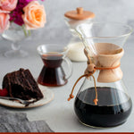 A Chemex Filter Drip Coffeemaker (CM-8A) filled with coffee sits on a table next to a glass mug, a slice of chocolate cake, and flowers, showcasing its elegant 40-ounce capacity.