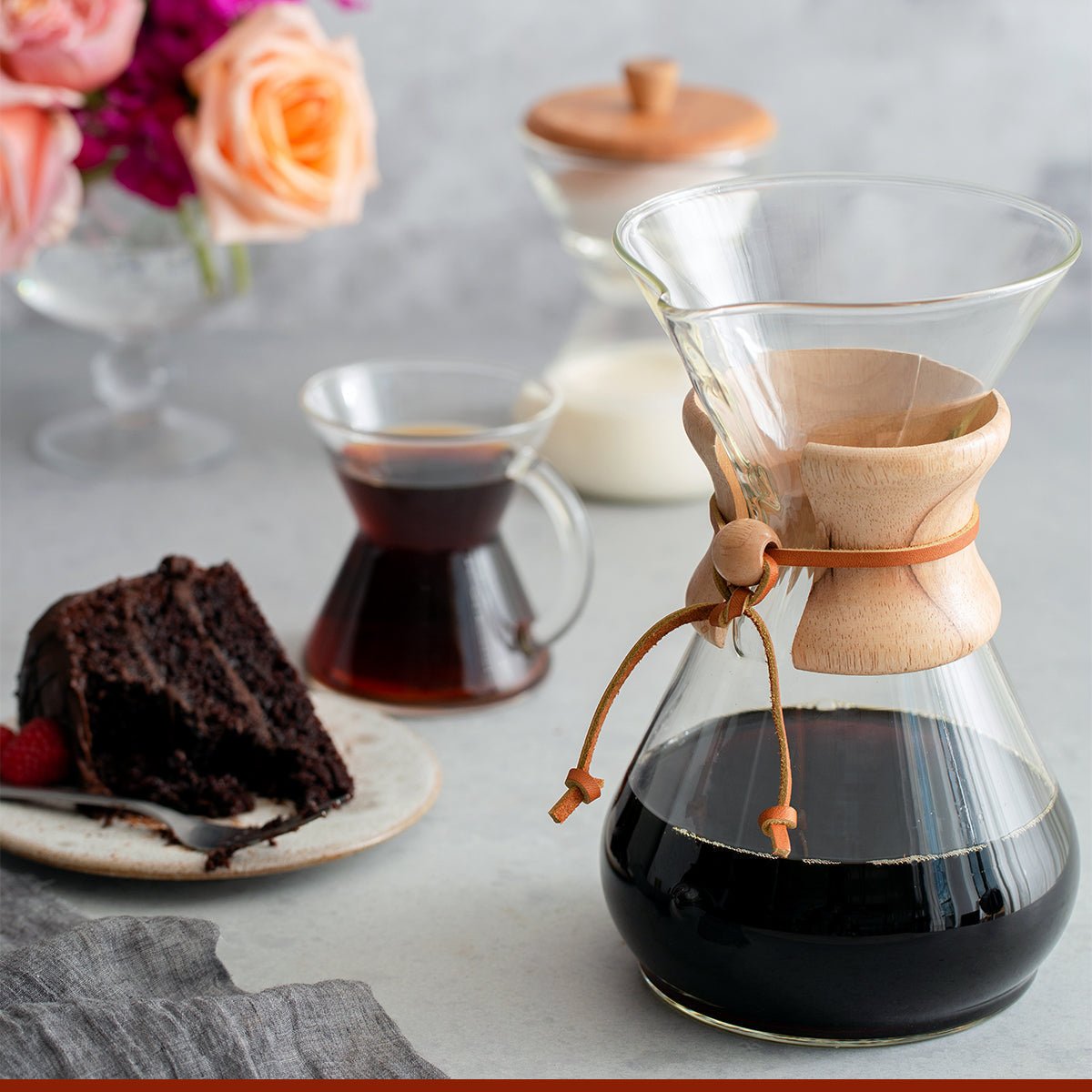 A Chemex Filter Drip Coffeemaker (CM-8A) sits on a table filled with coffee next to a glass cup, a plate with chocolate cake, and a vase of flowers in the background.