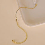 The JaxKelly Flat Chain Anklet is an 18k gold plated, tarnish-free anklet with a lobster clasp and extension links, offering a classic look. It's pictured on a beige surface near a light-colored tray for timeless style.