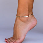 A close-up of an ankle showcases the JaxKelly Flat Chain Anklet, 18k gold plated and tarnish free. The toes have red nail polish against a plain light gray background.