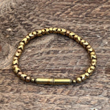 The Flint Beaded Single Bracelet by Tres Cuervos, featuring gold-toned magnetic beads and a cylindrical clasp, is displayed on a textured wooden surface.