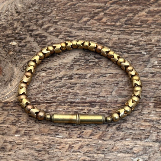 The Flint Beaded Single Bracelet by Tres Cuervos, featuring upcycled .22 shells and recycled glass beads with a cylindrical clasp, rests on a textured wooden surface.