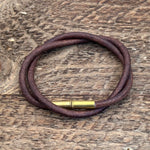 The Flint Bracelet by Tres Cuervos features upcycled .22 shells on a brown leather cord with a brass clasp, coiled on a textured wooden surface—ideal for those who appreciate rustic jewelry.