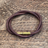 The Flint Bracelet by Tres Cuervos features a brown leather cord and brass clasp, coiled in a circle on a textured wooden surface.