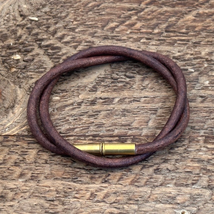 The Flint Bracelet by Tres Cuervos features a brown leather cord and brass clasp, coiled in a circle on a textured wooden surface.