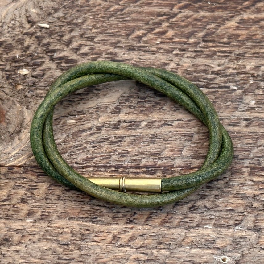 The Flint Bracelet by Tres Cuervos features a green leather cord and brass magnetic clasp, coiled into a circle and displayed on a textured wooden surface.