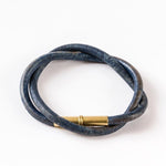The Flint Bracelet by Tres Cuervos features dark blue leather, a gold cylindrical magnetic clasp, and an oval coil design, displayed on a white background.