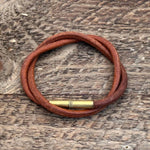 The Flint Bracelet by Tres Cuervos features a brown leather cord and a brass cylindrical clasp, artfully coiled on textured wood—a perfect rustic accessory.