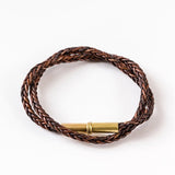 The Flint Braided Bracelet by Tres Cuervos features braided leather and a cylindrical gold magnetic clasp, shown coiled in a circle on a white background.